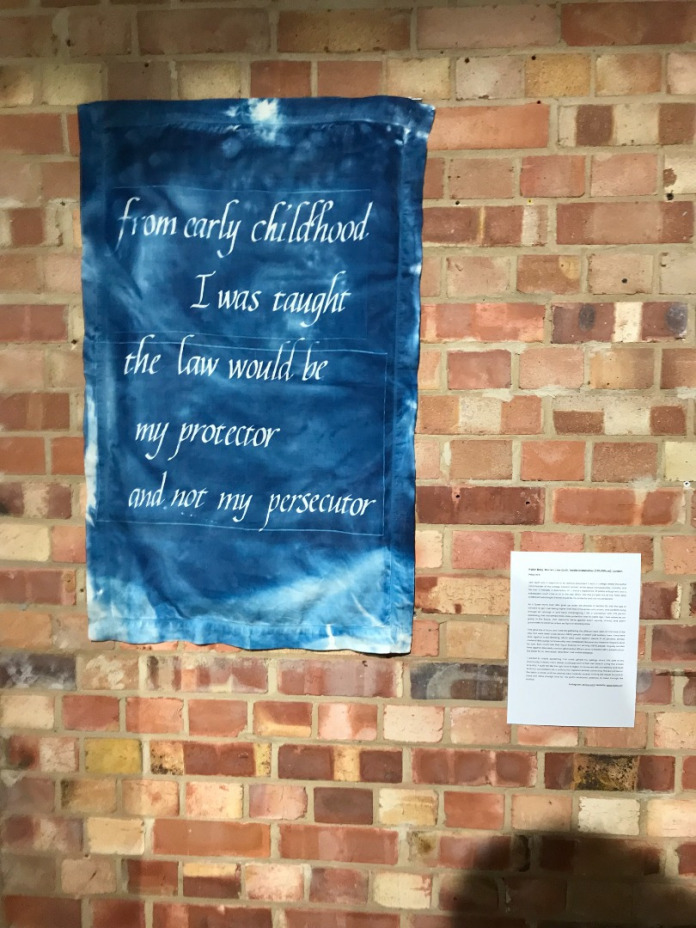 Title: Accompanying text to Law Quilt. A pillowcase hung vertically on a brick wall. The pillowcase is dyed a deep blue, with paler patches here and there. White text in neat hand lettering reads "from early childhood I was taught the law would be my protector and not my persecutor."