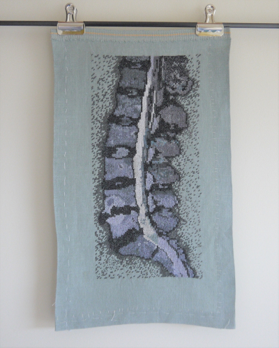 A piece of pale grey fabric clipped to a dowel, fixed against a wall. The fabric has an image sewn into it, made from hundreds of regularly placed, densely worked stitches, sewn in a grid. It's my lower spine taken from an MRI, looked at as a side-on slice so you can see the lordotic curve (which is mostly straight for me), and damage to several discs and two vertebrae.