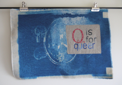 Q is for Queer. A piece of embroidered fabric is clipped to a dowel fastened to a wall. The fabric is dyed deep blue, and has images from a brain scan in pale blues and white. A small rectangle of light brown fabric is inset into the right side of the fabric, cutting into the largest brain scan image. The phrase “Q is for queer” is cross stitched into the brown fabric, the Q in pink, in a curly typeface, the other letters in a blocky sans serif font in shades of grey. 