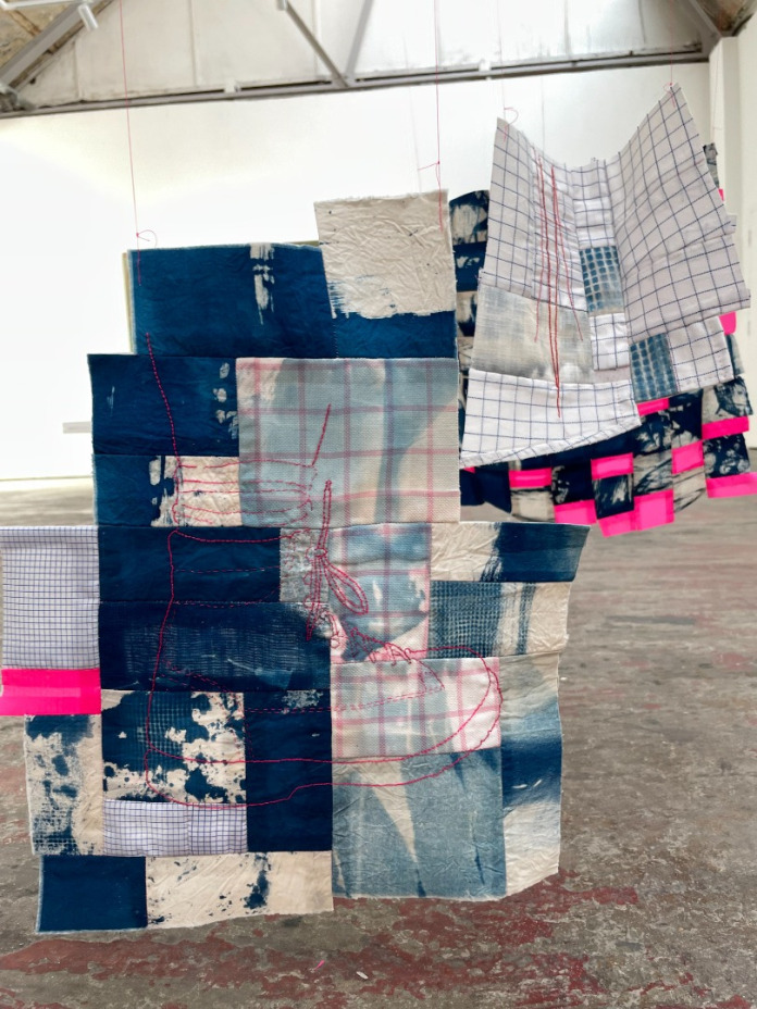 Title: Piecing Fragments, Boot. Detail of on of the patches from the larger hanging piece. An outline drawing of a boot and partial lower leg is sewn in fluorescent pink thread on the ground fabric, which is constructed from irregularly sized scraps of different shirt fabrics and fabric dyed deep blue. All the shirt fabric are either striped or checked with thin pink or blue lines. 