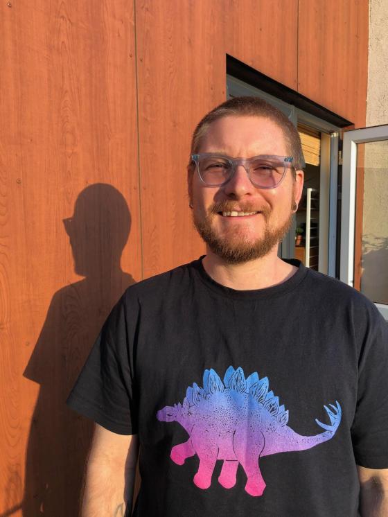 A photo of me standing in front of an external wooden-cladded wall. I'm squinting a bit in bright sunshine. I'm a white man in my late thirties, with very short light brown hair, clear blue ex-NHS specs, and a slightly scruffy beard & moustache. I'm wearing a black t-shirt with a stegosaur on. The stegosaur is a very chunky dinosaur on all fours, with a small head, long tail with four spikes on its tip, and a spine lined with angular plates. It's coloured blue along the spine plates, graduating through to pink at the toes. 