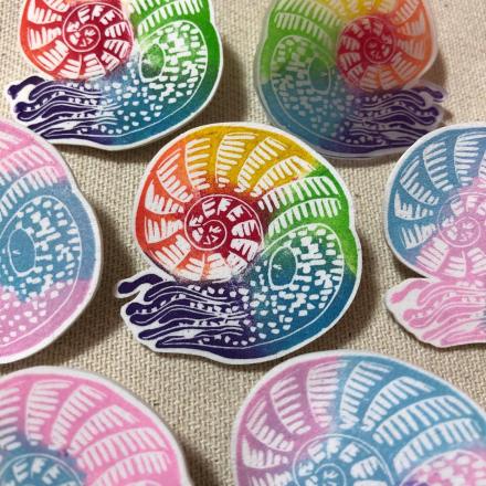 A cluster of 7 hand made badges. The badges are of ammonites, a prehistoric sea creature with a coiled shell, and a body ending in tentacles, like a squid crossed with a snail. The centre ammonite is rainbow coloured, and the surrounding ones are either rainbow coloured or are in pale pink & blue, like the trans pride flag.  