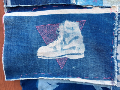 Queer Boot. A rectangle of fabric with an image dyed onto it, of an ink-drawn boot in shades of pale blue, on a dark blue background. Behind the boot is stitched hundreds of densely worked, small pink stitches, making a pink triangle, point facing downwards. 