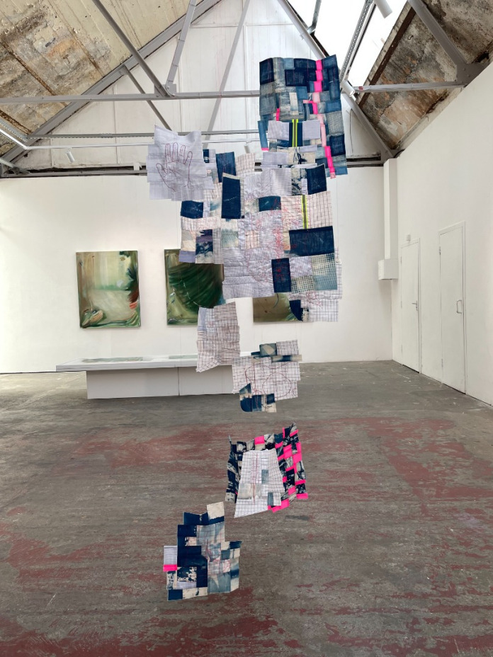 Title: Piecing Fragments. Artwork hanging in a gallery (an old single-storeuy warehouse building.) The piece is constructed of multiple fabric patches hung as a cluster from one of the ceiling beams. The pieces are made of patches of old shirt fabric, fluorescent pink fabric, and cyanotype dyed blue & whte linen and cotton. Large oil paintings in grey, green and brown tones are visible on the far wall.