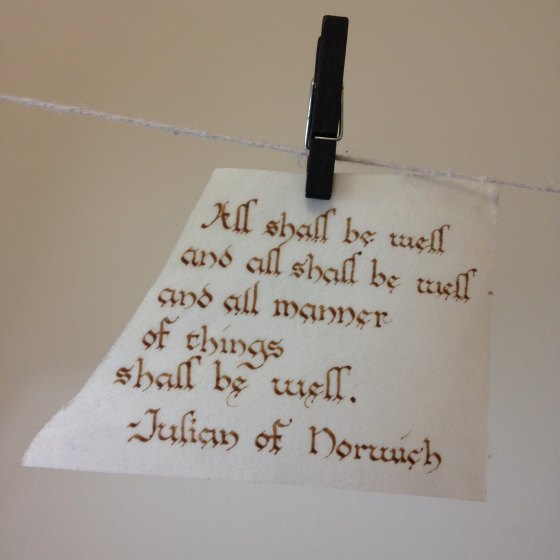 A scrap of parchment pegged to a string, with text written in a rounded gothic style reading "All shall be well, and all shall be well, and all manner of things shall be well" - Julian of Norwich