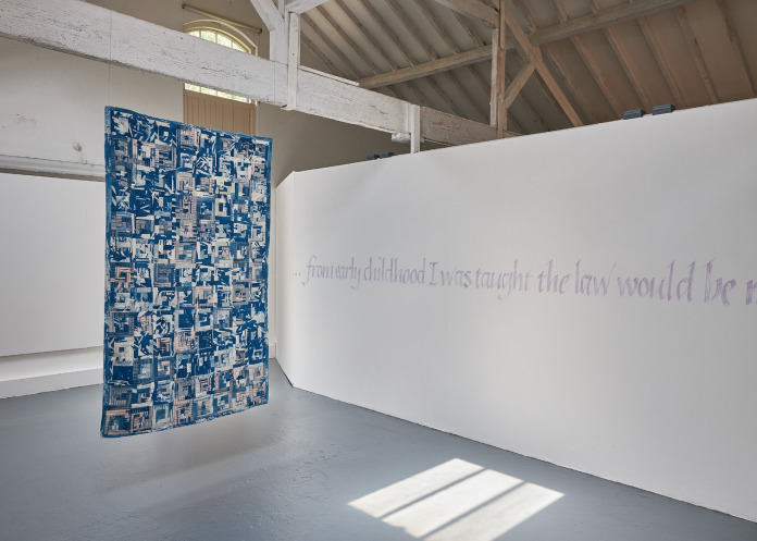 Title: Law Quilt. The quilt hangs from a beam in a white-walled room. The quilt's detail is unclear, from this distance it just has a random pattern of blue and white marks. Text painted on a nearby long wall reads "From early childhood I was taught the law would be..." the quote is cut off at the edge of the photo. 