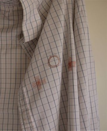 Mastectomy Shirt, detail. A formal cotton shirt with thin checked lines. Sewn on in thin deep pink silk is a circle where the nipple would be, a light pencil line visible out of alignment with the stitches. To the bottom right of the "nipple" is a small densely worked area of stitches marking the scar from  where one chest drain was placed. 