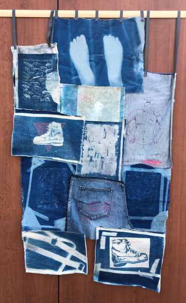 Masculinity Patchwork (Always In Progress). A fabric patchwork piece hanging from a wooden bar, against a wood panel external wall. The patchwork is constructed of irregular size and shape pieces of denim, most roughly rectangular, a couple taken from jeans, with the shape of the trouser panel. each piece has a different image dyed on in shades of blue. One has a sillhoutted pair of feet, this is the topmost piece. There's also a dildo harness down the bottom, a sewn self portrait of a man with scruffy beard and specs, a drawing of a clompy boot with a pink triangle sewn behind it, a page of a magazine with fish labelled "What is that queer fish?". It's a scruffy, patched together, wonkily constructed whole. 