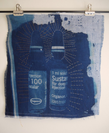 Holy Testosterone, Batman! An image printed on fabric in shades of blue dye, two glass ampoules with labels reading "Sustanon 100" and "Sustanon 250". Around the ampoules, long straight stitches in gold thread radiate outward like rays of sparkling light.