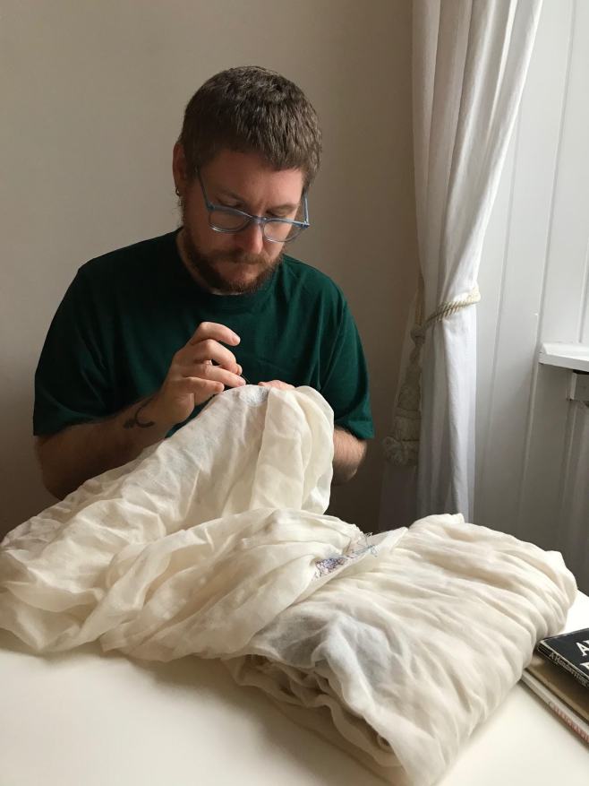 A photo of me sitting at a table, diffused light from the window on the right. I'm holding a draped piece of fine white fabric, one hand ready to pull a needle & thread through. I'm a white man in my late thirties, with short light brown hair, transparent blue specs, wearing a dark green t-shirt. 