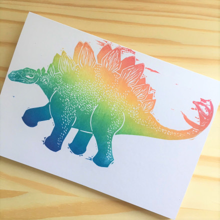 A card on a wooden board, the card has a picture of a stegosaur in rainbow colours printed on it. 