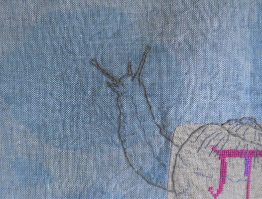 A close view of a piece of blue closely woven fabric, it has an inset piece of pale brown fabric set into it, and stitched across the seam where the two fabrics meet is a snail, shell over the brown fabric, grey body stretching up from the shell onto the blue fabric, eyes-on-stalks stretched upwards. Part of some cross stitch is just visible on the brown fabric, but you can't see enough to work out what it is. 